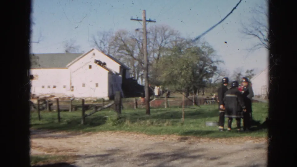 Firefighters in dark turnout gear stand near a grassy roadside with white farm buildings in Wauconda, Illinois, in 1968.