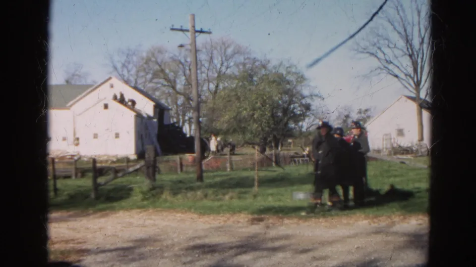 A home-movie view of firefighters gathered in grass near a white barn, utility pole, and bare trees in 1968 Wauconda.