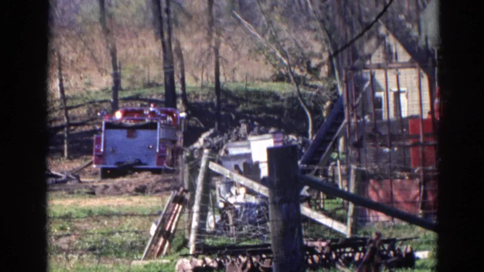 Red fire truck appears in a rural Wauconda yard, framed by dark film edges with fencing, scattered objects, and leafless trees visible.