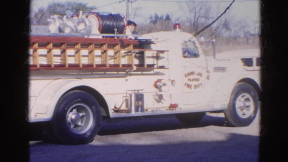 1968 Illinois home-movie frame showing a white volunteer fire department truck with ladder rack, hose reel, and door lettering.