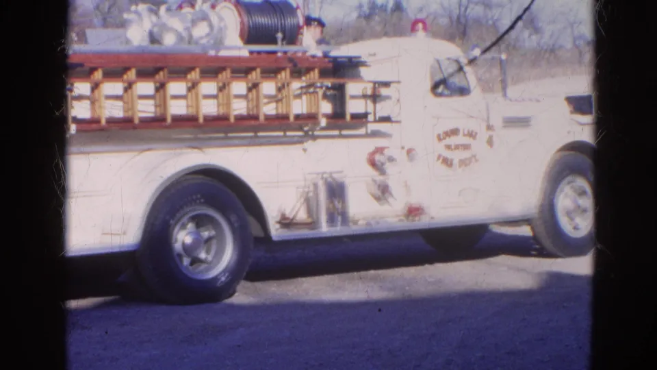 Home-movie frame from Wauconda area showing the firetruck side, ladder rack, rear wheel, and department markings.