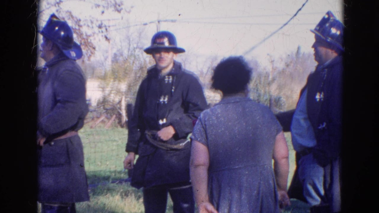 1968 Wauconda, Illinois 8mm frame showing firefighters in dark coats and helmets standing with a woman in a blue dress outdoors.
