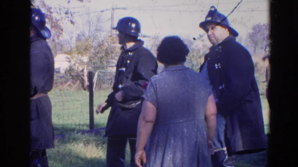 1968 Wauconda, Illinois frame with several firefighters in dark uniforms near a woman outdoors, with fencing and trees behind them.
