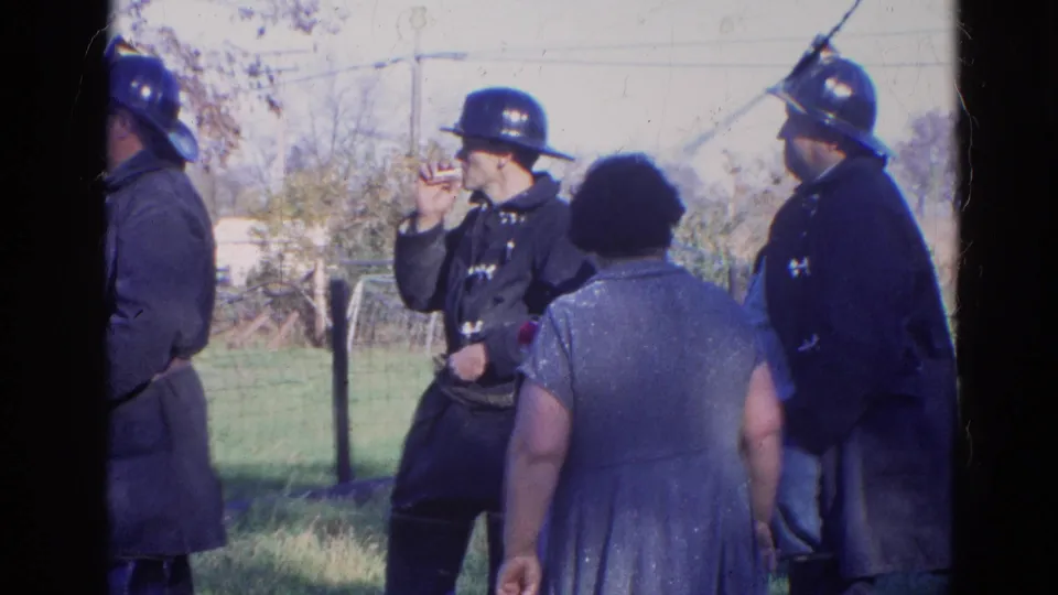 1968 Wauconda, Illinois 8mm frame showing a firefighter smoking during a pause beside colleagues and a civilian woman outdoors.