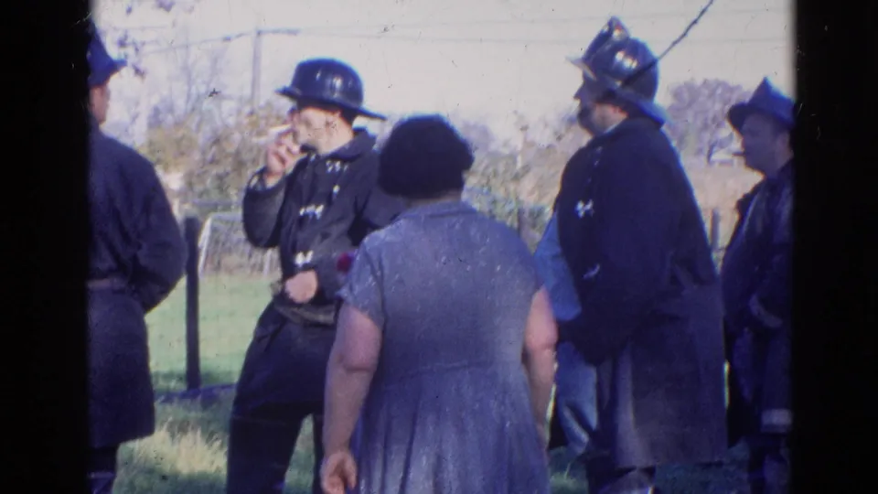 1968 Wauconda, Illinois frame of a firefighter smoking beside other helmeted firefighters and a woman in a blue dress outdoors.