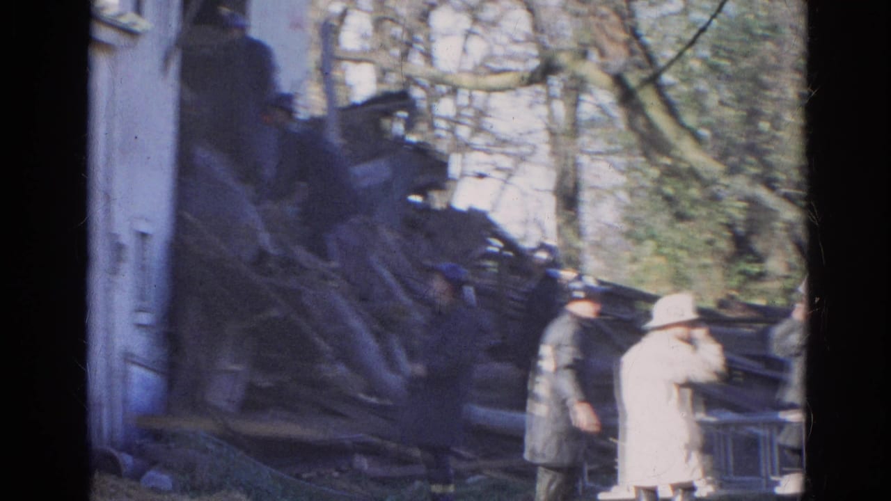 Firefighters and onlookers stand beside a collapsed wooden structure near a white building.