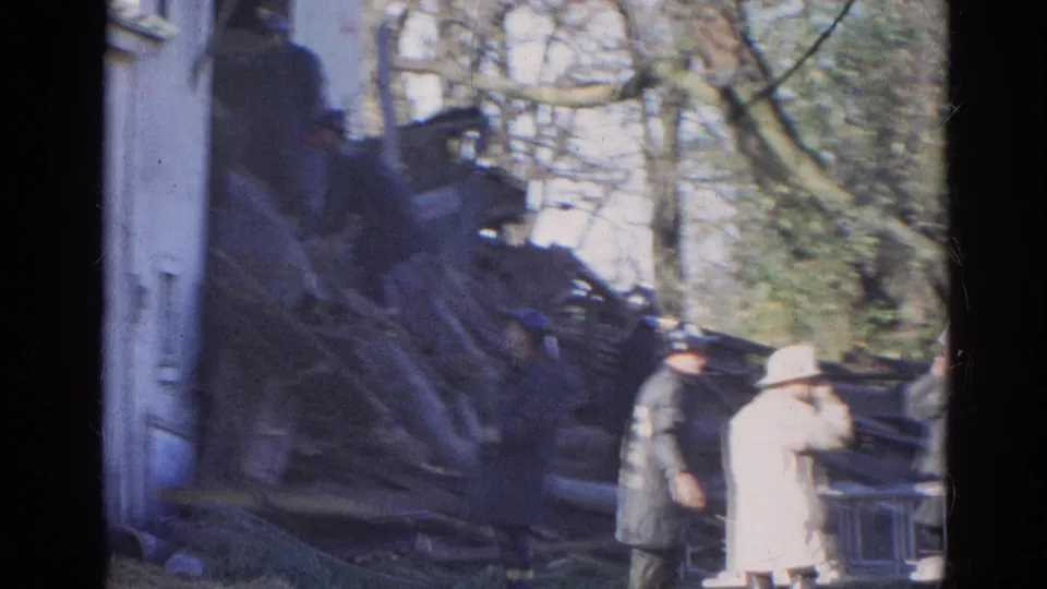 1968 Wauconda home-movie frame showing responders and a light-coated onlooker near collapsed wooden debris beside a white building.