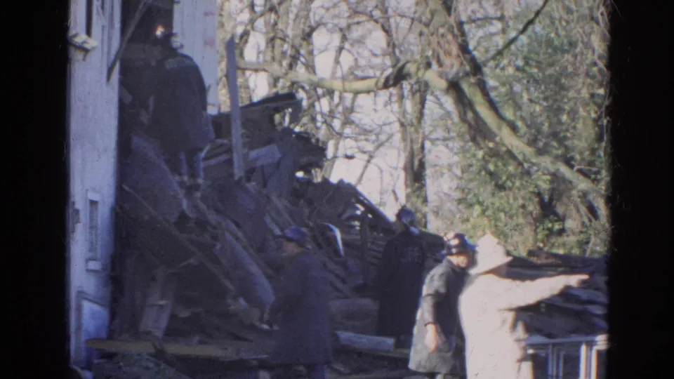 1968 Wauconda frame showing a firefighter helmet, dark-coated figures, and a light-coated onlooker pointing or reaching toward rubble.