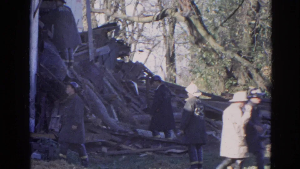 Final 1968 Wauconda frame with firefighters and onlookers near a large pile of structural debris beside a white building and winter trees.