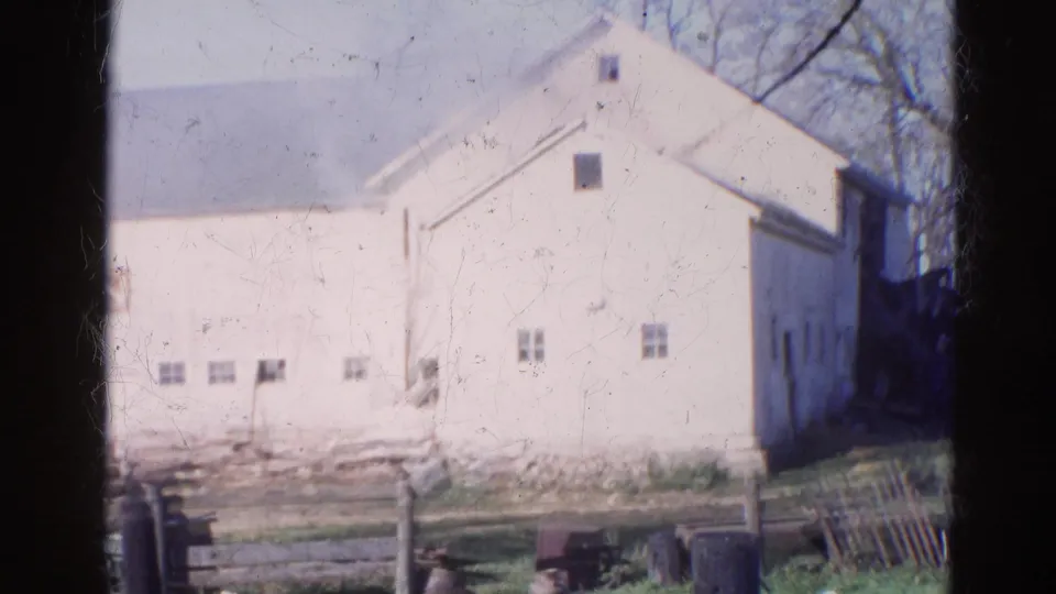 1968 Wauconda home-movie frame of a white farmhouse with small windows, debris, fencing, and trees under a bright sky.