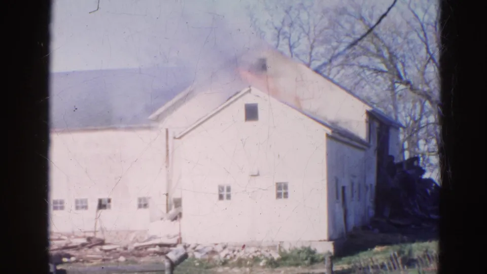 1968 Wauconda frame with visible smoke rising from the upper roof area of the white farmhouse, with trees and fencing nearby.