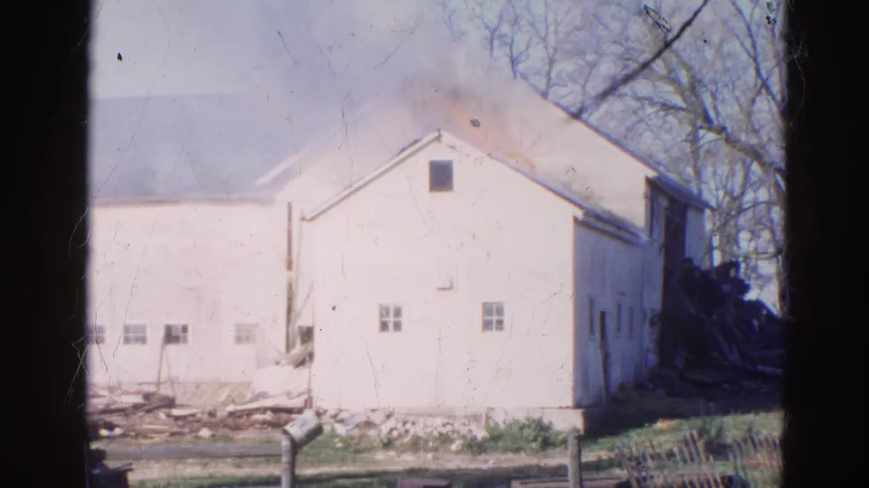 1968 Wauconda, Illinois 8mm frame of a white barn as smoke thickens near the roof and charred material appears beside the structure.