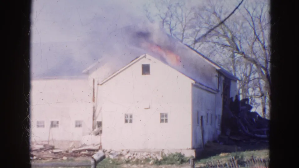 1968 Wauconda, Illinois 8mm frame with smoke and orange flame along the roof of a white barn, plus fence rails and debris in the foreground.