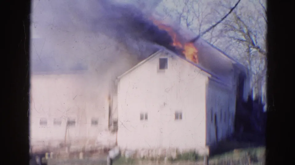 A white barn in Wauconda, Illinois burns in 1968, with orange flame at the roof edge and smoke spreading across the pale sky.