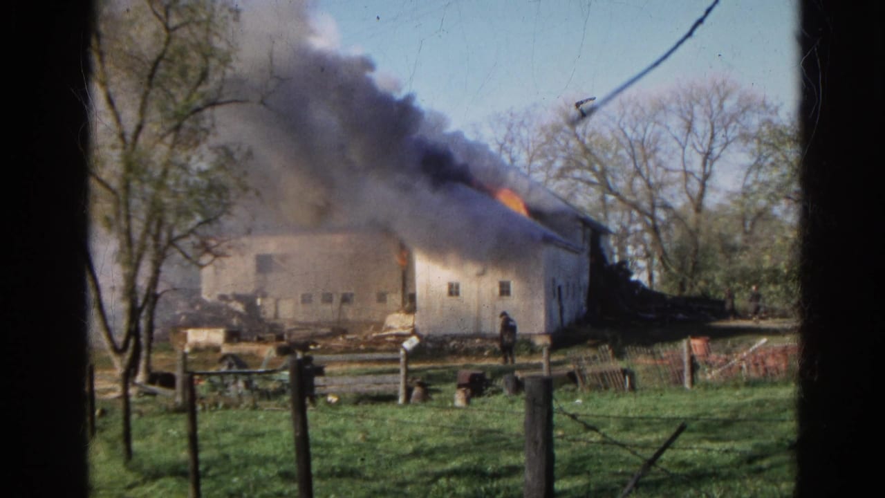 The 8mm view widens to show the burning white barn across grass, fence posts, bare trees, debris, and a plume of dark smoke.