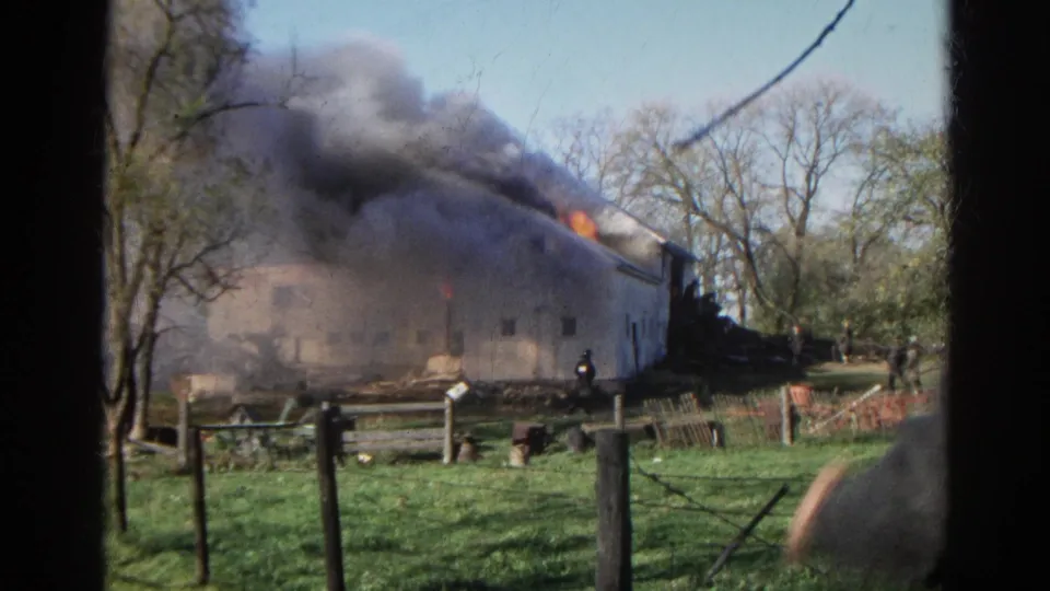A firefighter stands near the burning white barn as dark smoke pours from the roof and flames remain visible in the 1968 Wauconda footage.