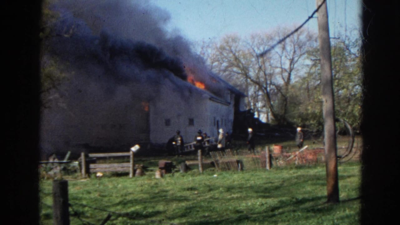 Barn-like building with roof flames and black smoke as firefighters stand near fences.