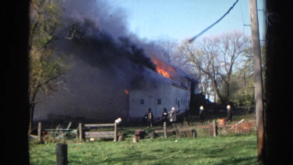 Wauconda, Illinois home-movie frame with firefighters below a burning barn-like building as smoke pours from the roof.