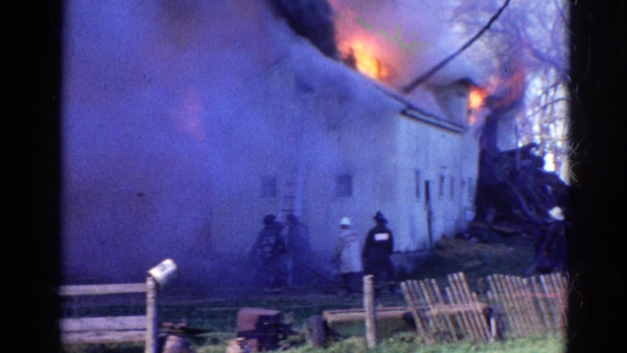 Firefighters stand near a burning building as smoke and flames rise behind a wooden fence.