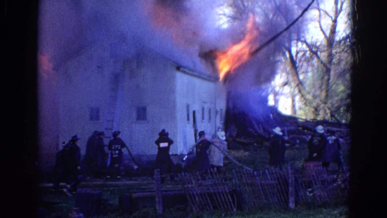 A group of firefighters stands near hoses as flames burn along the roof of a white building.