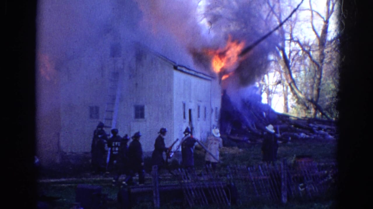 A 1968 home-movie frame shows firefighters, hoses, and a ladder beside a white rural building as fire burns near the roof.