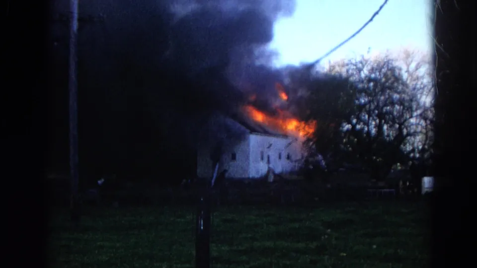 Wide 8mm view of the Wauconda house fire, with dark smoke, orange flames, a white building, trees, and a small figure in the foreground.