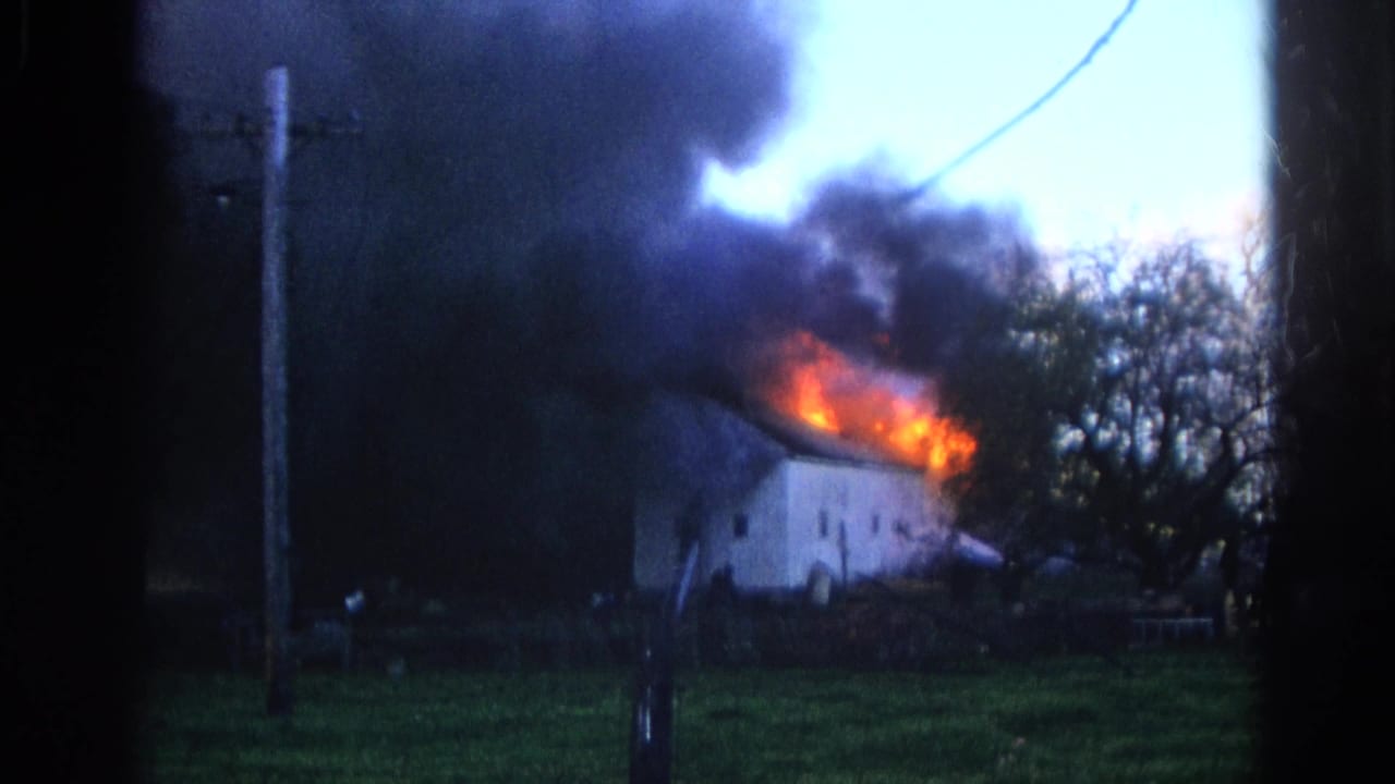 1968 Wauconda, Illinois 8mm frame showing a white house on fire, with flames along the roof and dark smoke rising behind a utility pole.