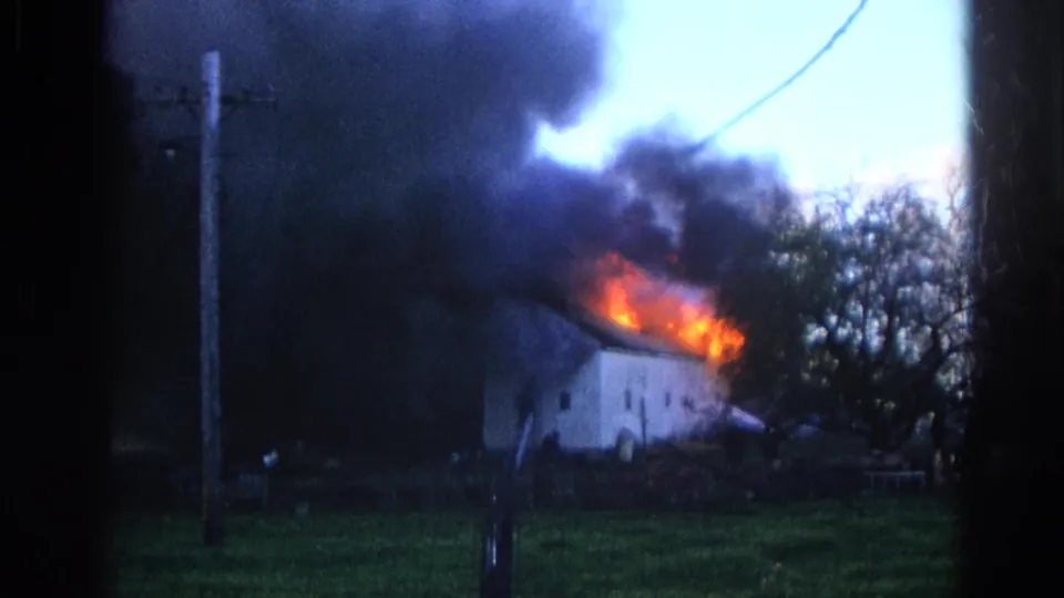 1968 Wauconda, Illinois 8mm frame showing a white house on fire, with flames along the roof and dark smoke rising behind a utility pole.