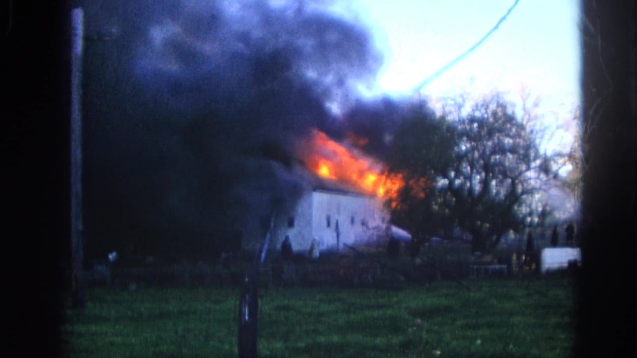 1968 Wauconda, Illinois 8mm frame of a white house engulfed at the roofline, with heavy smoke, bare trees, and a light vehicle or trailer at right.