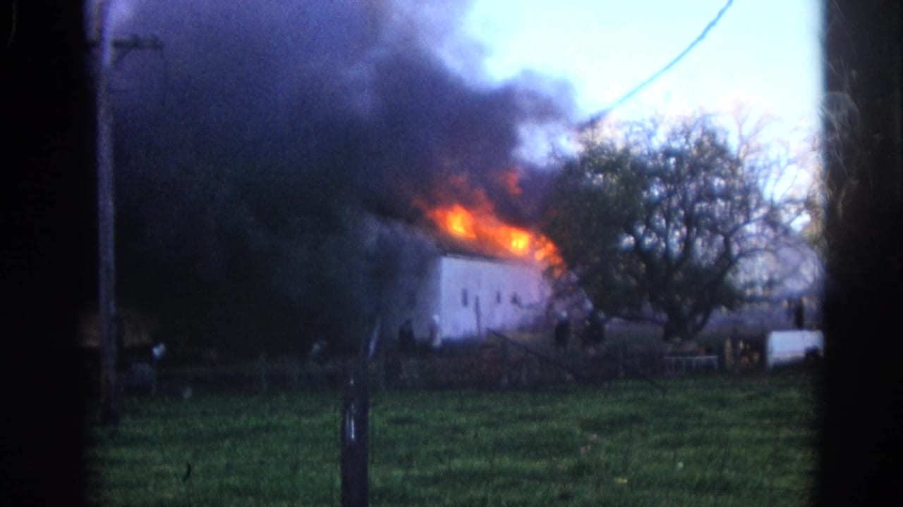 1968 Wauconda, Illinois 8mm frame showing a white structure on fire with dense smoke, orange flames, trees, and a grassy foreground.