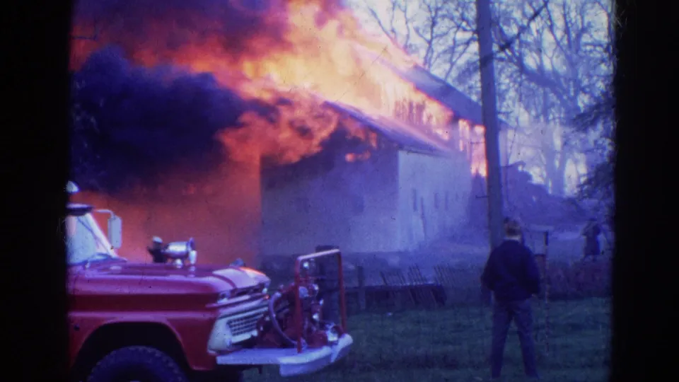 1968 Wauconda, Illinois frame showing a red fire truck, a standing person, hose equipment, flames, smoke, and the side of a burning building.