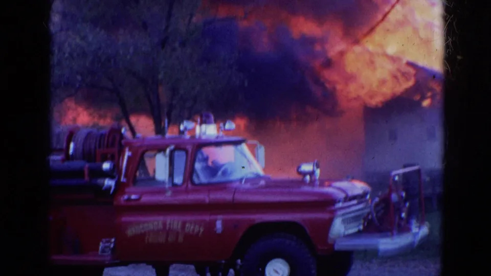 1968 Wauconda, Illinois frame with a red fire department truck in the foreground and a burning structure, flames, smoke, and responders nearby.