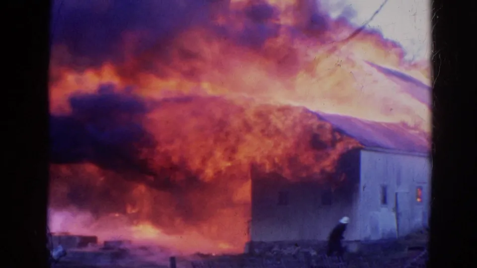 A firefighter bends near the side of a burning building in 1968 Wauconda footage, with intense flames behind the structure.