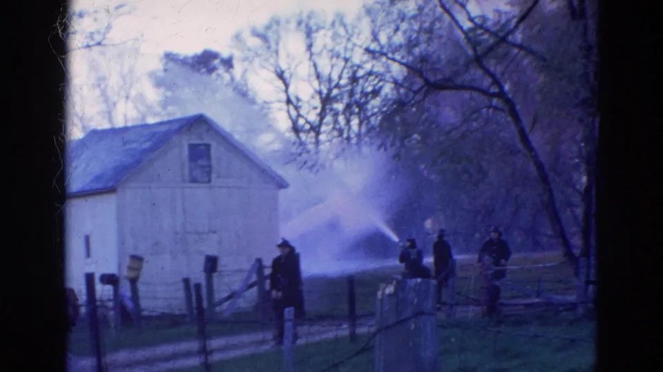 1968 Wauconda, Illinois frame showing firefighters with hoses near a light-colored rural building and smoke behind trees.