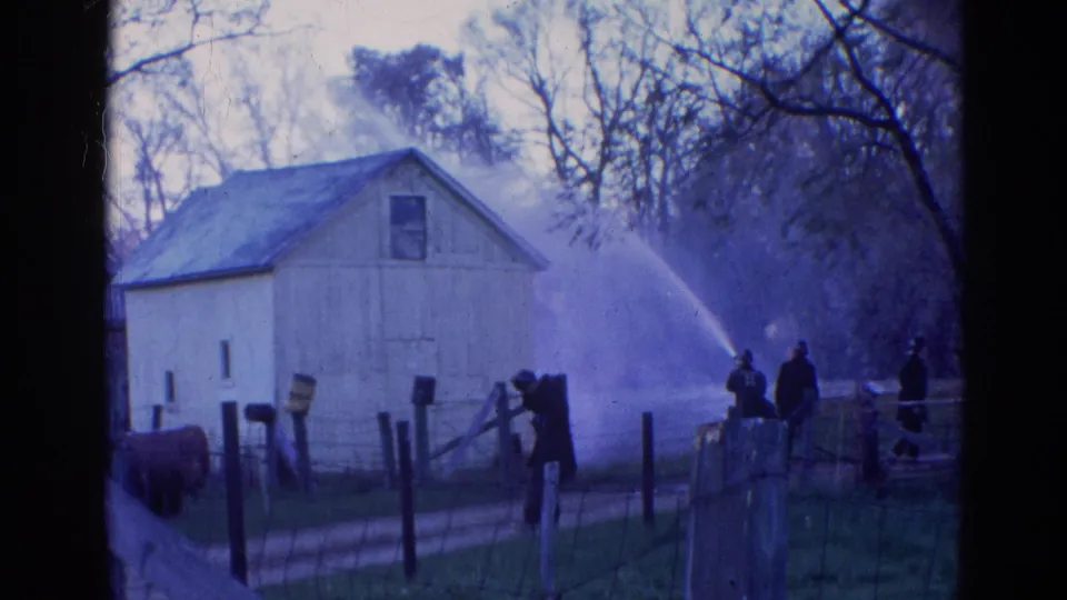 1968 Wauconda, Illinois 8mm frame with firefighters standing near a fence as water sprays beside a white barn.