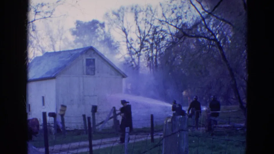 1968 Wauconda, Illinois 8mm frame showing firefighters using a hose across a rural fenced area beside a white barn.