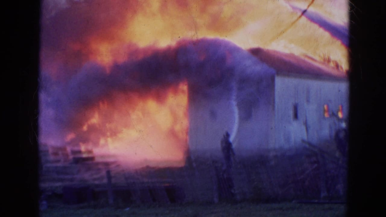 Large flames and black smoke surround a barn as a firefighter stands with a hose nearby.