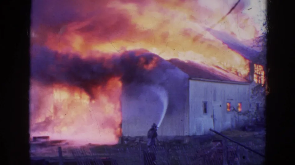 The 8mm frame shows a firefighter aiming a hose at a barn fire in Wauconda, Illinois, with dense smoke and flame behind the wall.
