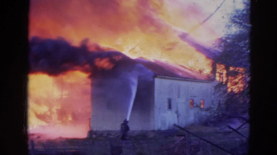 Vintage 8mm view of a light-colored building amid heavy flames, with a firefighter and hose visible in the foreground.