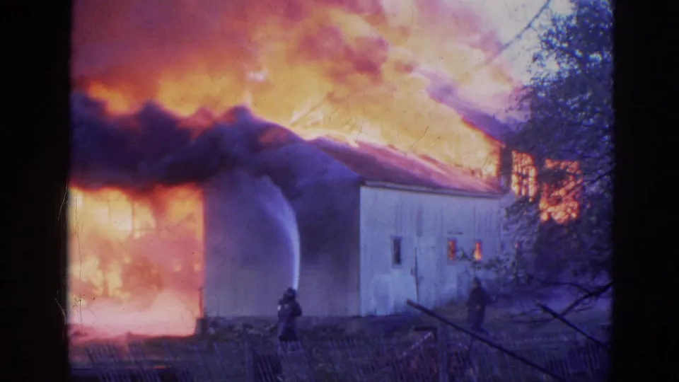 1968 Wauconda fire scene with firefighters near a building wall as water streams into dense smoke and bright flames.
