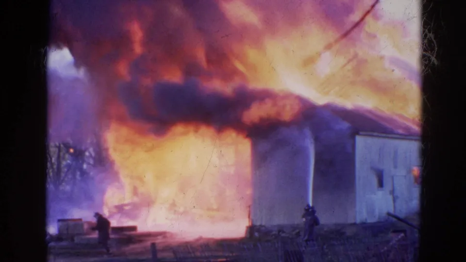 Handheld 8mm view of a Wauconda structure fire with firefighters silhouetted against bright flames in 1968.