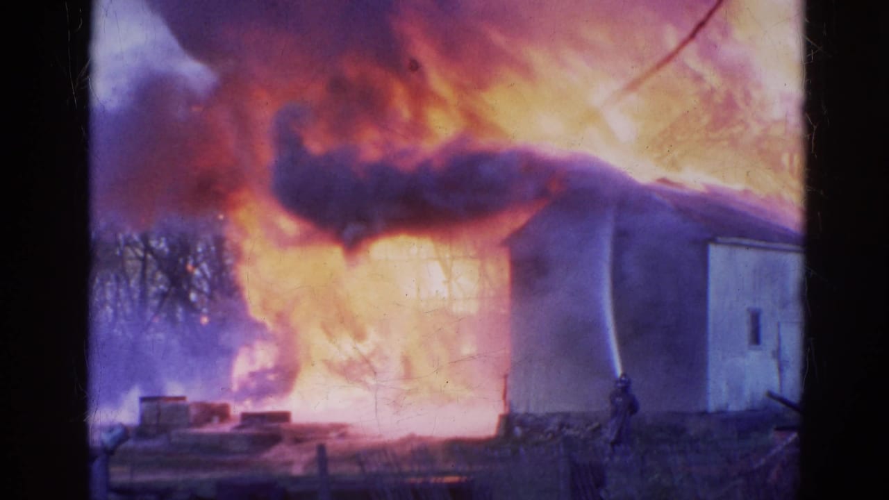 Large flames and dark smoke engulf a light-colored building as a firefighter stands nearby.