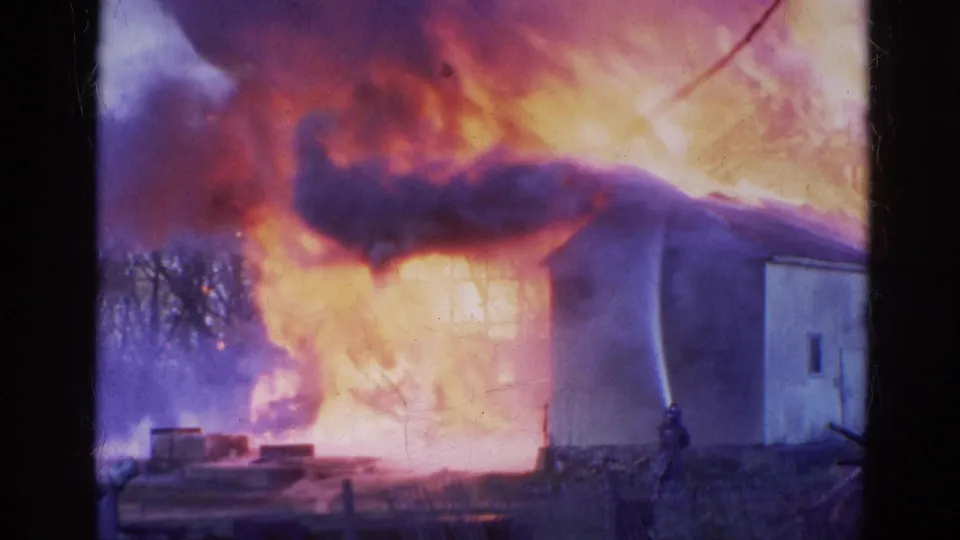 1968 Wauconda, Illinois 8mm frame showing a light-colored building consumed by bright flames, heavy smoke, and a distant firefighter silhouette.
