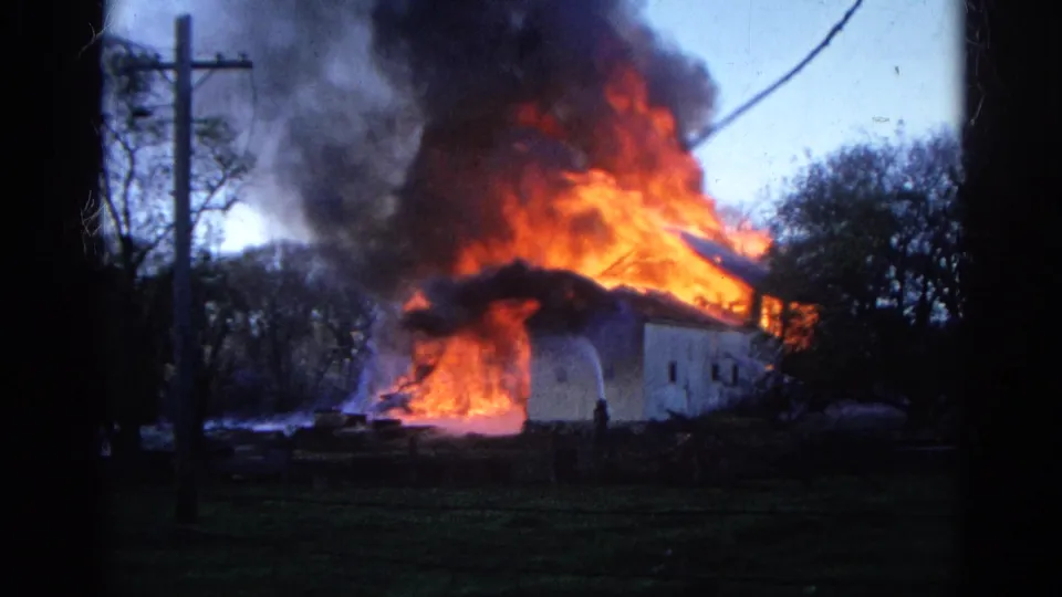 1968 Wauconda, Illinois frame showing a light-colored structure burning intensely, with smoke overhead and a distant person in the foreground.