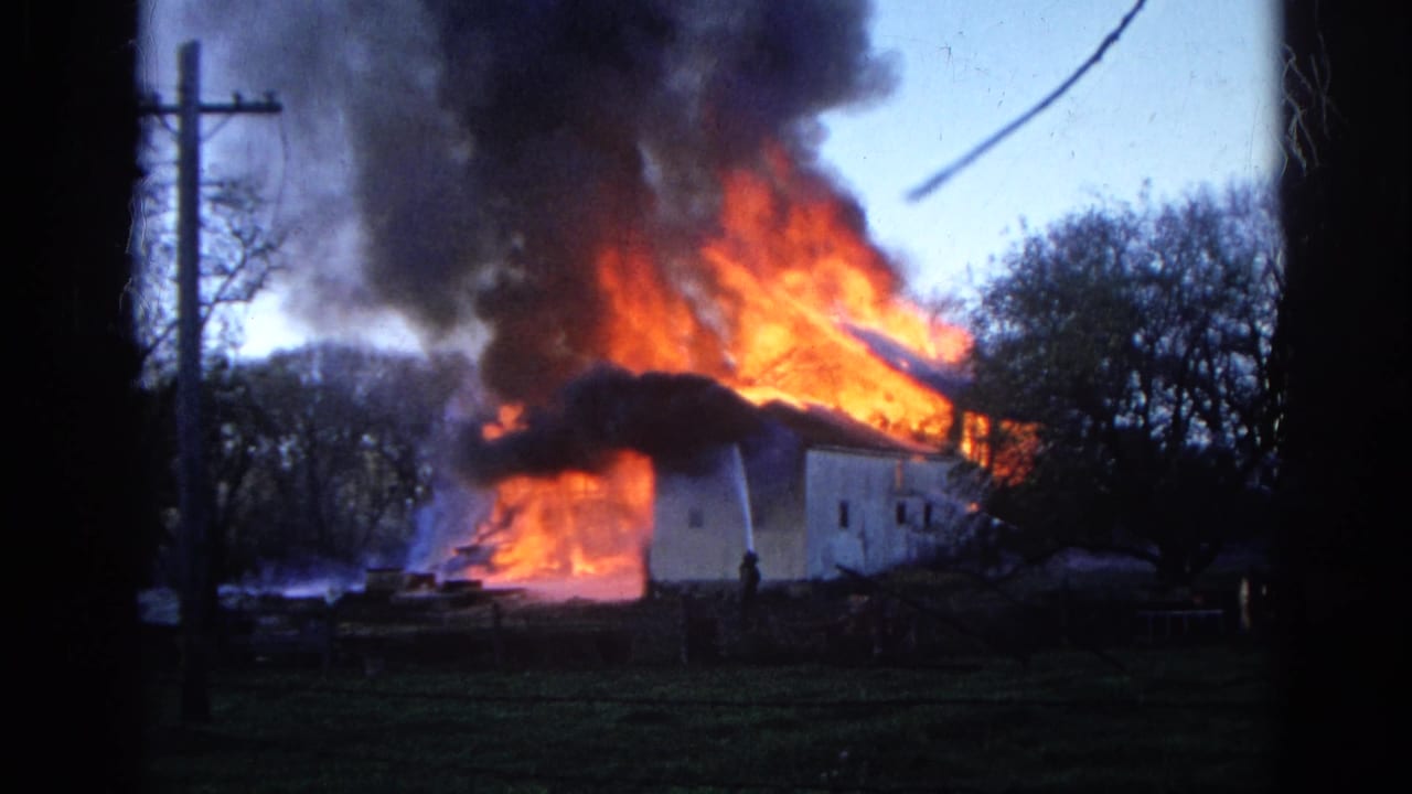 White rural building burning with orange flames and black smoke behind a utility pole.