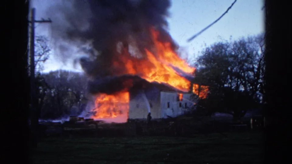 Wauconda, Illinois home-movie frame showing flames consuming the roof area of a white rural building with dark smoke above.