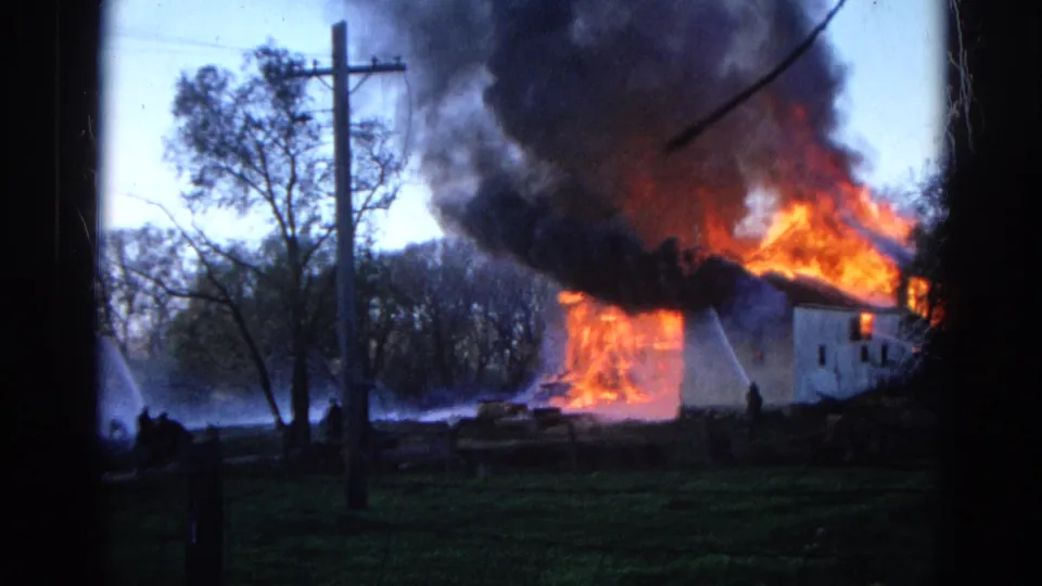 Wauconda, Illinois 1968 frame with silhouetted people and water spray at left while a white rural building burns under heavy smoke.