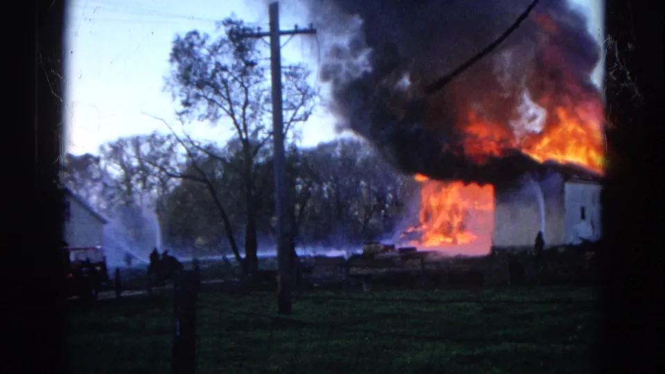 1968 Wauconda, Illinois 8mm frame showing a white house engulfed in flames, black smoke overhead, and water streams aimed at the fire.