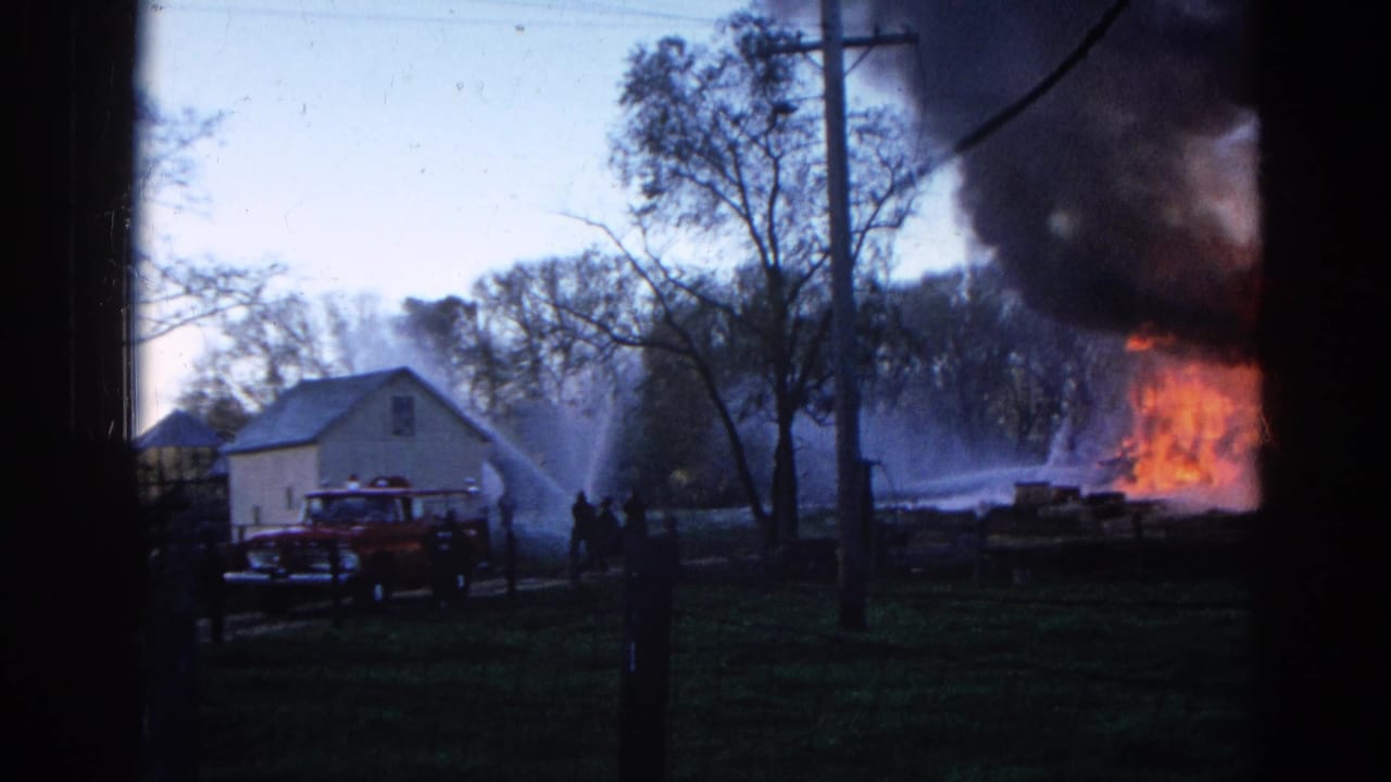 Firefighters spray water near a house as flames and dark smoke rise beside it.