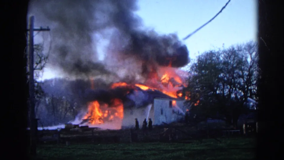 1968 Wauconda, Illinois frame of a house fire with orange flames, heavy smoke, hose spray, trees, and distant people.